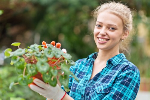 A young woman with blonde hair tied back, wearing a blue plaid shirt and gardening gloves, is standing in a well-maintained garden and holding a small potted plant. The garden features a lush, green lawn with densely grown grass, bordered by a neat hedge which appears to be regularly trimmed. In the background, there are mature trees and flowering shrubs, indicating a landscaped outdoor space typical of suburban gardens in Ruislip. The scene is outdoors under bright natural daylight, likely in mild weather conditions suitable for gardening, and the garden surface showcases a mix of grass, soil, and paved pathways. This setting reflects typical gardening and landscaping services such as hedge trimming and plant care, offered by companies like Hedge Trimming Ruislip, serving local homeowners seeking professional outdoor maintenance and garden enhancement.