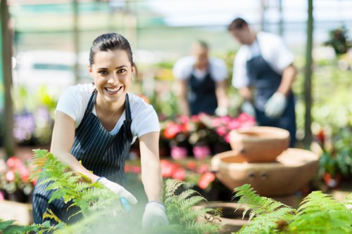 A smiling woman wearing a striped apron and disposable gloves is working in a brightly lit greenhouse or plant nursery, leaning over a bed of lush green ferns with feathery fronds. Behind her, other gardening staff are visible, wearing aprons and engaged in plants and flower arrangement tasks. The environment features a variety of potted plants, floral displays, and decorative stone water features, suggesting a professional gardening or landscaping service setting in Ruislip. The greenhouse has a transparent roof and glass walls, allowing natural light to illuminate the vibrant green foliage and colourful flowers, which provide a natural contrast to the earthy soil and garden beds. The scene demonstrates outdoor garden maintenance, planting activity, and plant care, aligning with the services provided by Hedge Trimming Ruislip in local gardening and landscaping works, including plant health and growth management in a controlled environment.