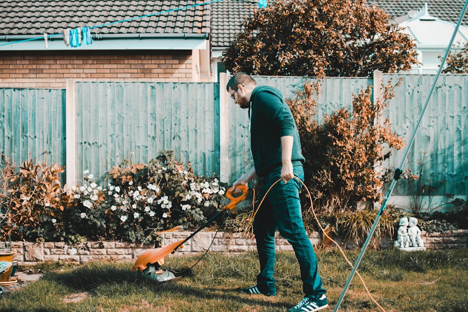 A man with short dark hair, wearing glasses, a dark hoodie, and blue jeans, is trimming a well-maintained garden lawn using an electric grass trimmer. The grass appears freshly cut, and there are patc