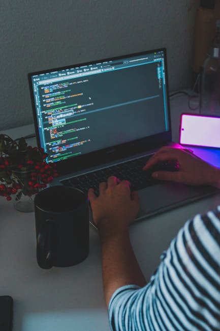 A close-up view of a person's hands working on a laptop computer displaying a coding interface with colorful lines of code. The workspace includes a black mug positioned to the left of the laptop, a s